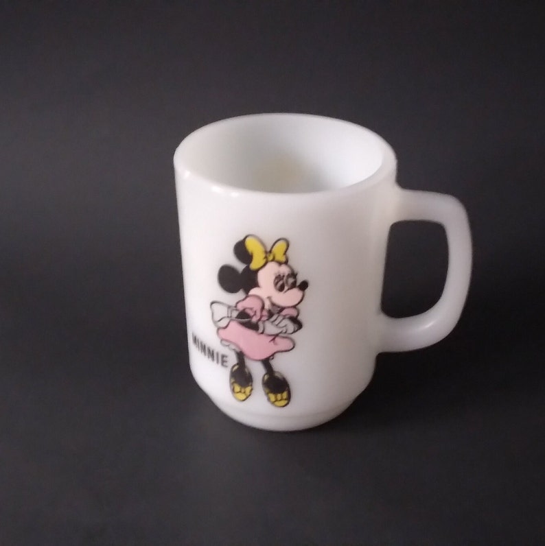 May include: White ceramic mug with a cartoon image of Minnie Mouse wearing a pink dress and yellow shoes. The mug has the word "Minnie" printed on the side.