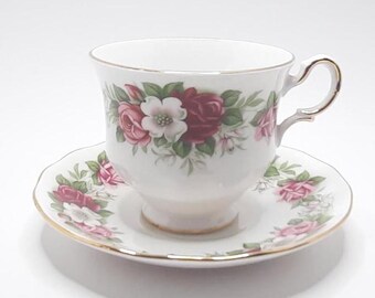May include: A white teacup and saucer set with a floral pattern featuring pink and red roses, white flowers, and green leaves. The set has a gold trim around the edges.