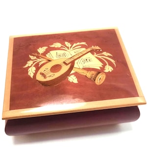 May include: A wooden music box with a decorative inlaid design. The top features a lute, musical notes, and a horn, set against a reddish-brown background. The box has a light-colored border and a dark purple base.