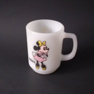 May include: White ceramic mug with a cartoon image of Minnie Mouse wearing a pink dress and yellow bow. The mug has a black handle and the word "MINNIE" is printed on the bottom.