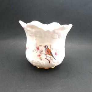 Puede incluir: Un jarrón de cerámica blanca con borde festoneado. El jarrón presenta un diseño pintado de un pájaro posado en una rama con flores rosas. La base tiene un ribete dorado.