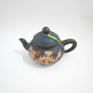May include: A small, dark blue teapot with a dragon design. The teapot has a green string tied around the lid.