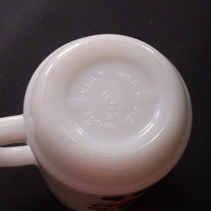 May include: White ceramic mug with a red and black design. The bottom of the mug has the text "Made in USA" and "Anchor Hocking".