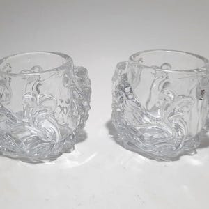 May include: Two clear glass votive candle holders. Each has a round opening and is decorated with a floral design. The sides of the holders have a textured, sculpted appearance. The candle holders are sitting on a white surface.