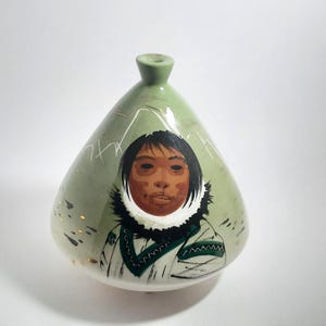 May include: A green ceramic vase with a painted portrait of a person in traditional Inuit clothing. The vase has a rounded shape with a narrow neck and is decorated with white mountain outlines and gold accents. The person's face is centered on the vase.