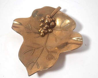 May include: A vintage brass ashtray in the shape of a leaf, featuring a detailed vein pattern and a cluster of grapes. The ashtray has a smooth, polished finish and is designed with a small lip for holding cigarettes.
