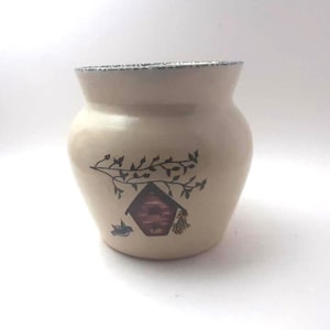 May include: A beige ceramic jar with a birdhouse and bird design. The jar has a wide opening and a slightly curved body. The birdhouse is red and brown, with a branch and leaves design. The rim has a blue and white pattern.