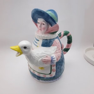May include: A ceramic teapot shaped like a woman in a blue and green hat holding a white goose. The teapot has a pink and blue floral pattern.