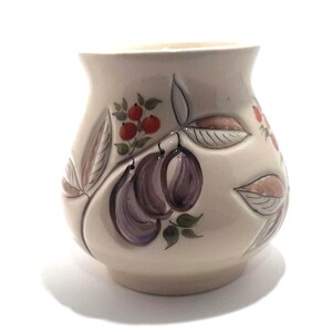 May include: A cream-colored ceramic vase with a rounded body and a slightly flared rim. The vase is decorated with hand-painted illustrations of purple plums, red berries, and green leaves. The design is outlined in a darker color.