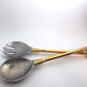 Op de afbeelding: Een saladeservies bestaande uit een vork en een lepel. De vork heeft drie tanden en de lepel heeft een afgeronde kom. Beide hebben zilverkleurige koppen en goudkleurige handvatten met een geribbeld ontwerp bij de kop.