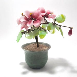 May include: A decorative glass flower arrangement in a small, light green ceramic pot. The arrangement features pink flowers, green leaves, and a brown stem. The pot is filled with small, beige pebbles, creating a miniature bonsai tree.