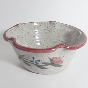 Puede incluir: Un pequeño cuenco de cerámica blanca con un borde rosa y un diseño floral. El cuenco tiene un borde festoneado y está decorado con una flor rosa, hojas azules y tallos verdes.