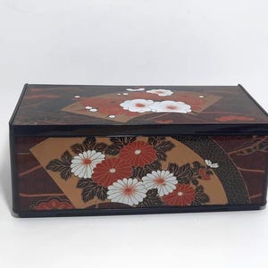 Puede incluir: Una caja rectangular marrón oscuro con un borde negro. La caja está decorada con un diseño floral con flores blancas y rojas sobre un fondo beige. La tapa y la parte delantera de la caja tienen el mismo diseño.