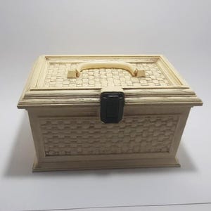 Peut inclure: Une boîte de rangement rectangulaire beige avec un motif tissé sur les côtés et le dessus. Elle est dotée d'une poignée incurvée et d'un loquet noir. La boîte est faite d'un matériau de couleur claire et repose sur une surface blanche.