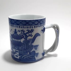 May include: White ceramic mug with a blue Willow pattern design. The mug features a twisted handle and a detailed landscape scene with a pagoda, trees, and a bridge. The rim has a blue geometric pattern.