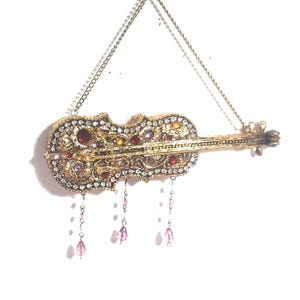 May include: Ornate gold-colored violin-shaped ornament, embellished with sparkling clear, red, and purple gemstones. The decorative piece hangs from a delicate chain and features dangling pink beads. A detailed, vintage-style design.