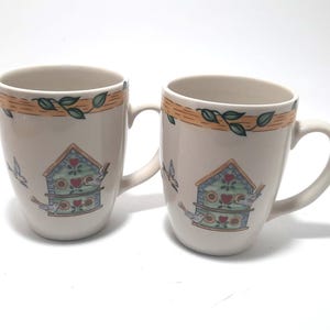 May include: Two off-white ceramic mugs with a birdhouse design. Each mug features a birdhouse with red hearts, green leaves, and flying birds. The top border has a brown and orange design with green leaves. The mugs have a curved handle.
