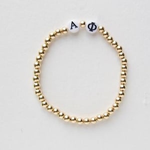 May include: A gold beaded bracelet with white letter beads spelling out "A" and "Φ" in navy blue. The bracelet is made of small, round, gold-colored beads.