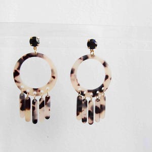 May include: A pair of dangle earrings with a cream and brown tortoiseshell pattern. Each earring features a black gem at the top, a circular hoop, and several rectangular dangles. The earrings are set against a white background.
