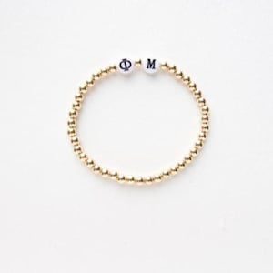 May include: Gold beaded bracelet with white letter beads spelling out "Φ M". The bracelet is made of small, round, gold-colored beads strung together. The white letter beads are centered on the bracelet.