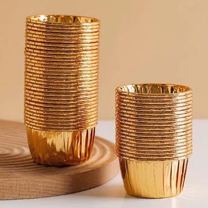 May include: A stack of gold foil cupcake cases, with a smaller stack of the same cases in front of them. The cases are made of a shiny, metallic material and have a fluted design.