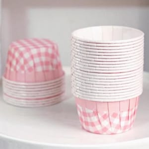 May include: A stack of white and pink gingham cupcake cases. The cases are on a white tray, with another stack in the background. The cupcake cases are for baking and are made of paper.