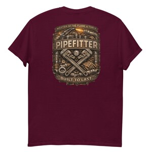 以下が含まれることがあります： バーガンディ色のTシャツ。詳細なグラフィックには「PIPEFITTER」、「MASTER OF THE FLAME & TORCH」、「BUILT TO LAST」の文字が含まれています。交差したレンチ、パイプ、その他のツールと炎の効果が特徴です。