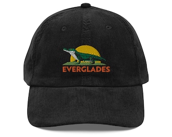 May include: Black corduroy baseball cap with an embroidered design. The design features a green alligator, a yellow sun, and the word "EVERGLADES" in orange. The cap has a curved peak and a button on top.