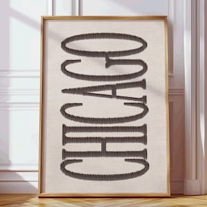 Può includere: Un'opera d'arte incorniciata con la parola "CHICAGO" in stile ricamato verticale. Il testo marrone scuro si staglia su uno sfondo beige, racchiuso in una cornice di legno marrone chiaro. L'opera è presentata contro un muro bianco.