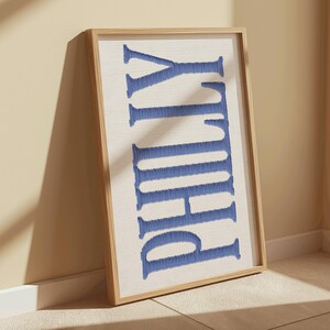 May include: A framed artwork featuring the word "PHILLY" in blue embroidered letters against a cream-colored background. The wooden frame complements the artwork, leaning against a beige wall and floor.