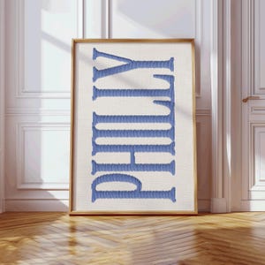 May include: A framed print featuring the word "PHILLY" in vertical, blue embroidered letters. The letters are stitched on a cream-colored background, encased in a wooden frame. The artwork is displayed against a white wall and wooden floor.