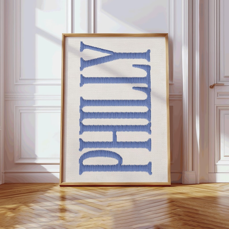 May include: A framed print featuring the word "PHILLY" in vertical, blue embroidered letters. The letters are stitched on a cream-colored background, encased in a wooden frame. The artwork is displayed against a white wall and wooden floor.