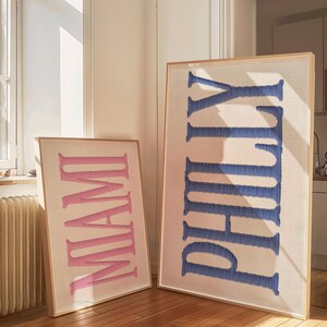 May include: Two framed wall art pieces. One displays the word "MIAMI" in pink, the other "PHILLY" in blue. Both are embroidered on a neutral fabric and set in light wood frames. The art pieces are leaning against a wall on a hardwood floor.