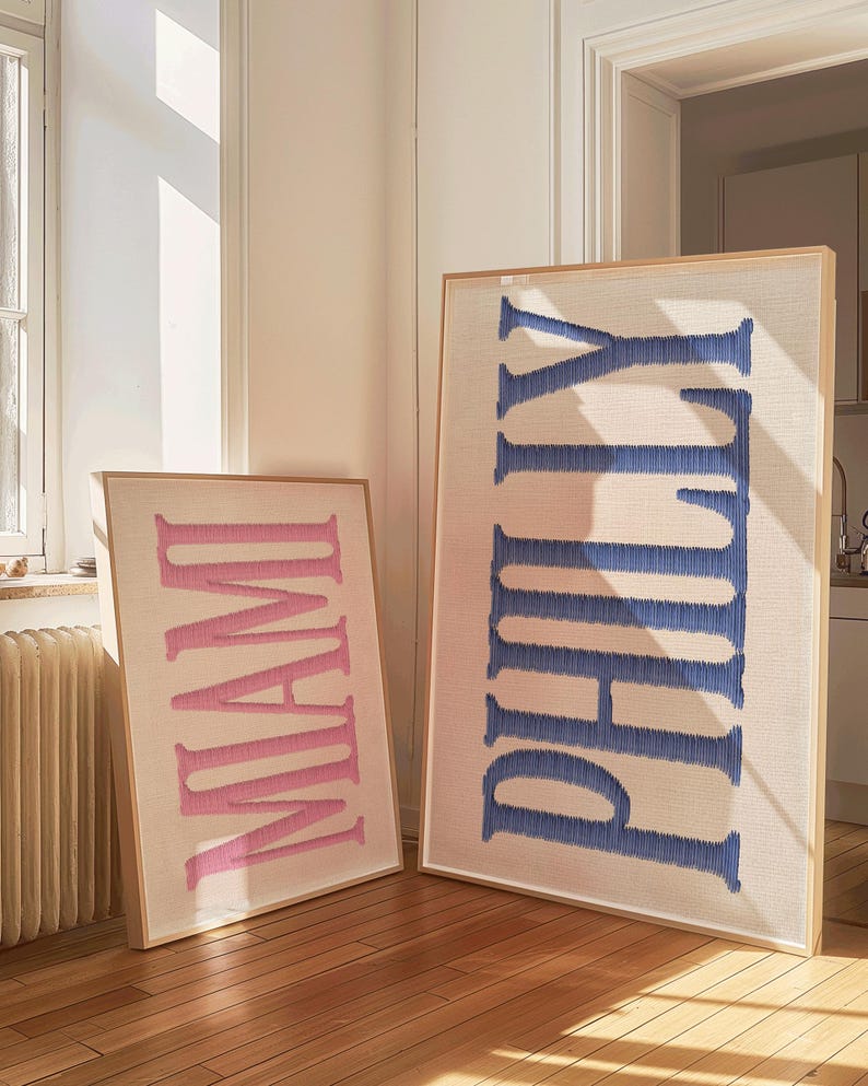 May include: Two framed wall art pieces. One displays the word "MIAMI" in pink, the other "PHILLY" in blue. Both are embroidered on a neutral fabric and set in light wood frames. The art pieces are leaning against a wall on a hardwood floor.