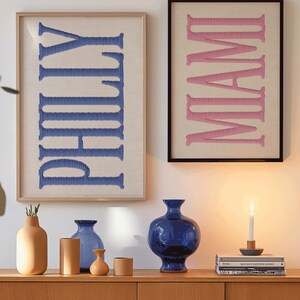 May include: Two framed wall art pieces with embroidered text. One reads "PHILLY" in blue on a beige background, the other "MIAMI" in pink. Below, a wooden cabinet displays various vases in shades of blue and tan, and a lit candle.