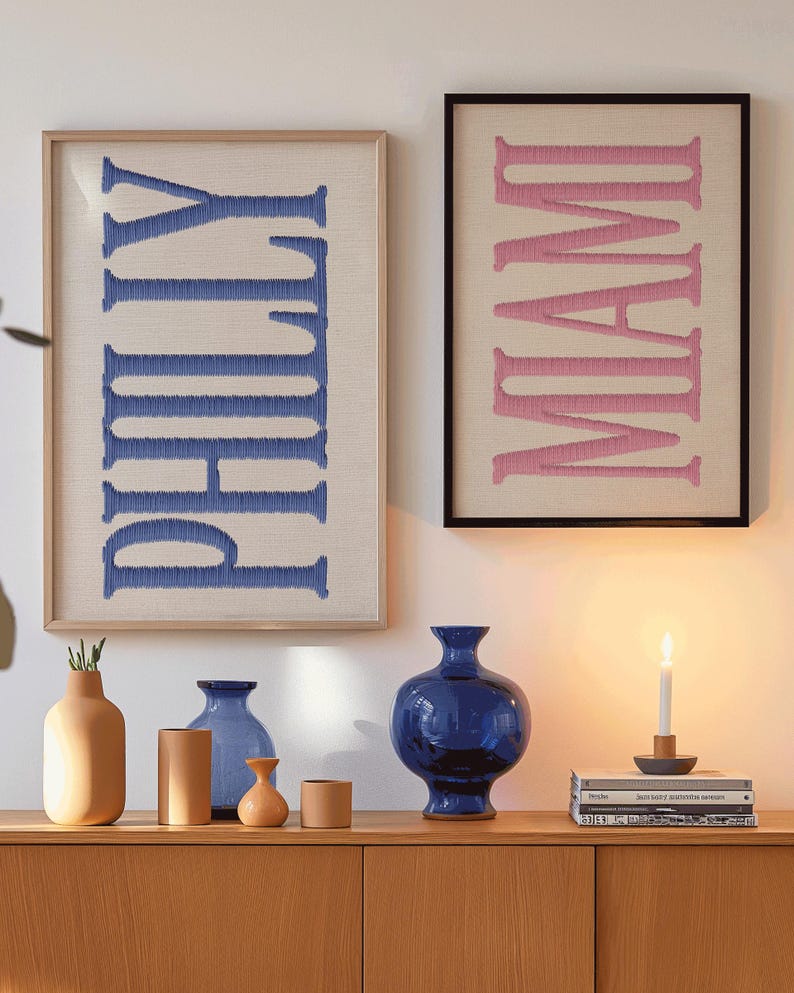 May include: Two framed wall art pieces with embroidered text. One reads "PHILLY" in blue on a beige background, the other "MIAMI" in pink. Below, a wooden cabinet displays various vases in shades of blue and tan, and a lit candle.
