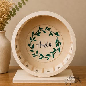 May include: A wooden hamster wheel with a light beige finish. The wheel features a central circular design with the name "Amon" in black script, surrounded by a green leaf wreath. The wheel is mounted on a rectangular base.