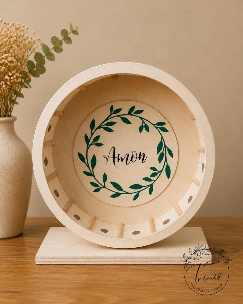 May include: A wooden hamster wheel with a light beige finish. The wheel features a central circular design with the name "Amon" in black script, surrounded by a green leaf wreath. The wheel is mounted on a rectangular base.