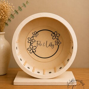 May include: A wooden hamster wheel with a light-colored finish. The wheel features a floral design with the text "Feli & Lilly" in the center. The wheel is mounted on a rectangular wooden base. The shop's logo is in the bottom right corner.