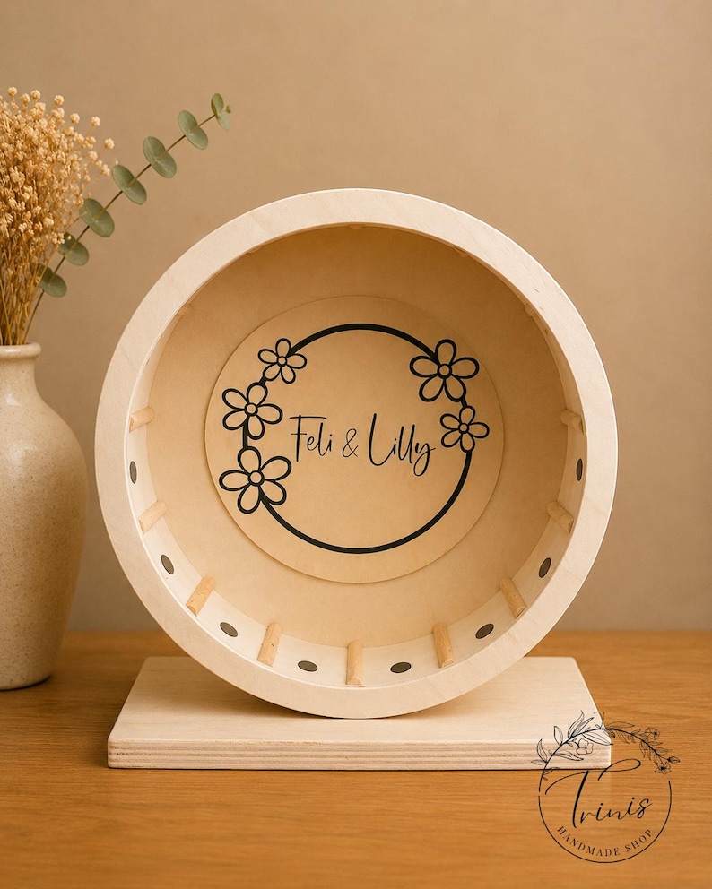 May include: A wooden hamster wheel with a light-colored finish. The wheel features a floral design with the text "Feli & Lilly" in the center. The wheel is mounted on a rectangular wooden base. The shop's logo is in the bottom right corner.