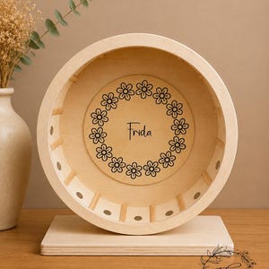 May include: A wooden hamster wheel with a light finish. The wheel has a circular design with a floral wreath and the name "Frida" in the center. The wheel is mounted on a rectangular base. The Trinis Handmade Shop logo is in the bottom right corner.