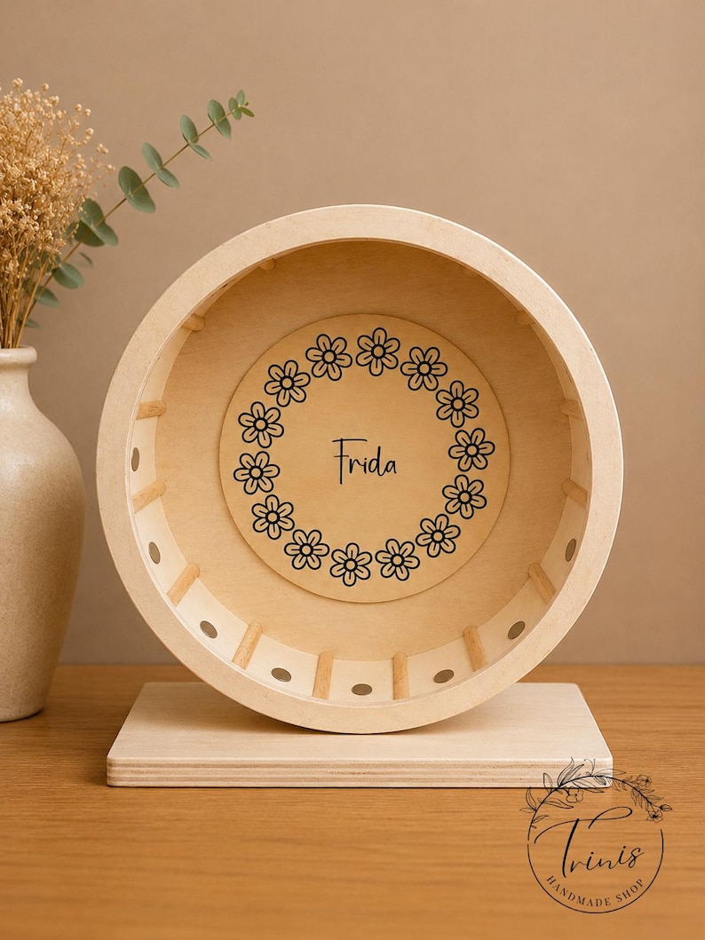 May include: A wooden hamster wheel with a light finish. The wheel has a circular design with a floral wreath and the name "Frida" in the center. The wheel is mounted on a rectangular base. The Trinis Handmade Shop logo is in the bottom right corner.