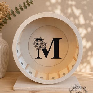May include: A wooden decorative wheel with the letter "M" in black, adorned with a floral design. The wheel is set on a wooden base. The item is a handmade product.