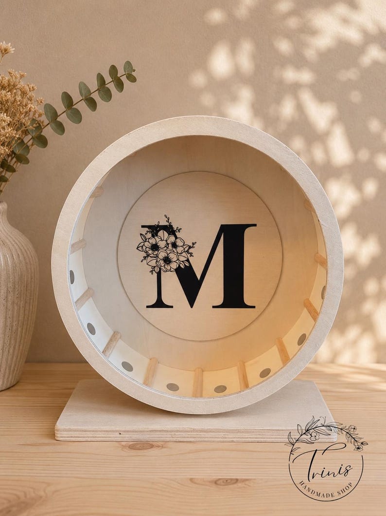 May include: A wooden decorative wheel with the letter "M" in black, adorned with a floral design. The wheel is set on a wooden base. The item is a handmade product.