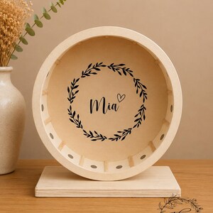 May include: A wooden hamster wheel with a light-colored finish. The wheel features the name "Mia" in black script, encircled by a black floral wreath. The wheel is mounted on a rectangular base, and the background is a neutral beige.