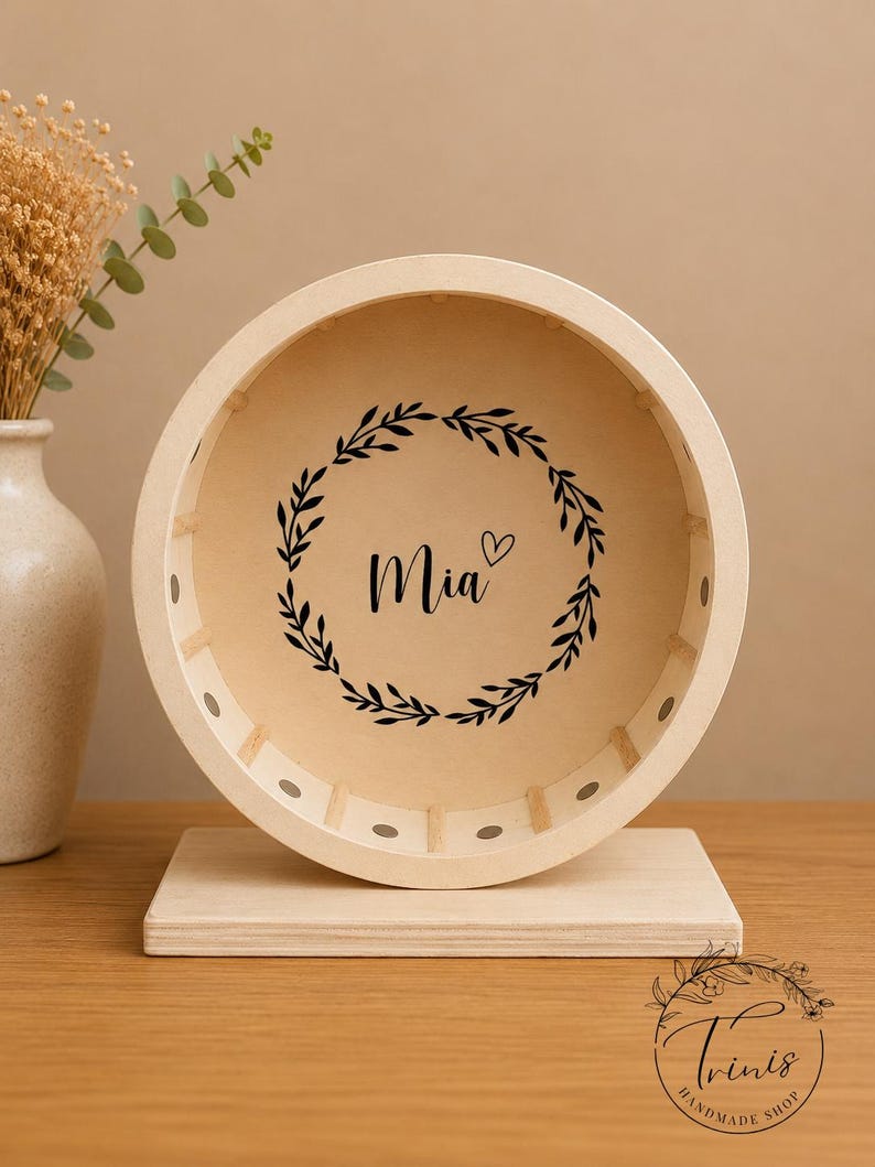 May include: A wooden hamster wheel with a light-colored finish. The wheel features the name "Mia" in black script, encircled by a black floral wreath. The wheel is mounted on a rectangular base, and the background is a neutral beige.