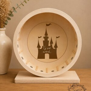 May include: A wooden hamster wheel with a castle silhouette and the text "Es war einmal..." in brown. The wheel is set on a rectangular base, with a light wood finish. The background includes a vase with dried flowers.