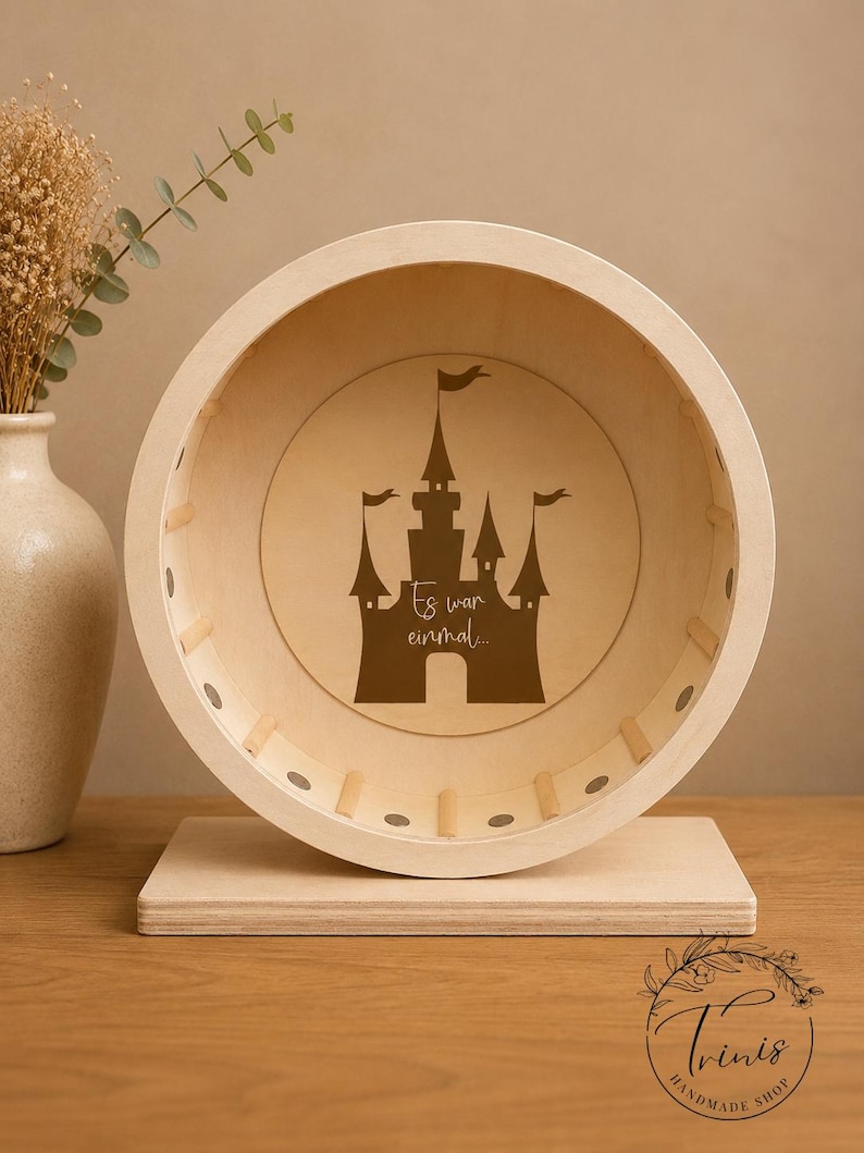 May include: A wooden hamster wheel with a castle silhouette and the text "Es war einmal..." in brown. The wheel is set on a rectangular base, with a light wood finish. The background includes a vase with dried flowers.