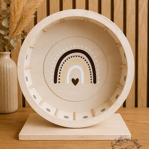 May include: A wooden hamster wheel with a rainbow design in the center. The wheel is beige with a light brown rainbow and a heart. The wheel is on a wooden base. The background is a wooden wall.