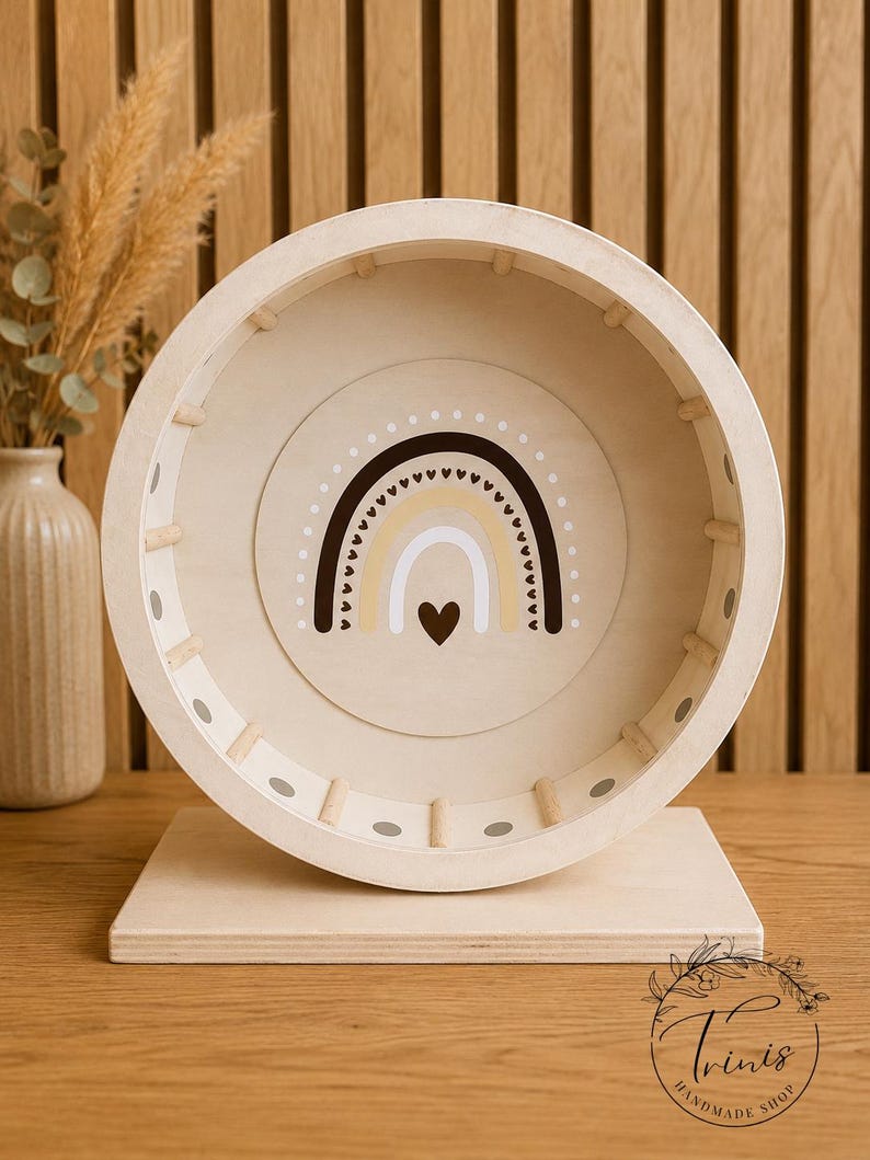 May include: A wooden hamster wheel with a rainbow design in the center. The wheel is beige with a light brown rainbow and a heart. The wheel is on a wooden base. The background is a wooden wall.
